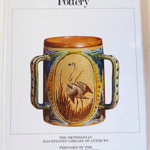 May include: A brown and yellow ceramic mug with two handles and a detailed design of a bird in a field. The mug is titled "Pottery" and is part of "The Smithsonian Illustrated Library of Antiques" prepared by the "Cooper-Hewitt Museum".