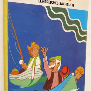 May include: A vintage children's book cover with a blue background and green waves. The title is "der Fluß" in orange letters. The subtitle is "LEHRREICHES SACHBUCH" in black letters. The cover features three cartoon characters in boats on the water. One character is fishing with a long brown fishing rod.