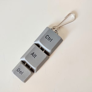 Puede incluir: Un llavero con tres teclas de plástico gris etiquetadas como "Ctrl", "Alt" y "Del".