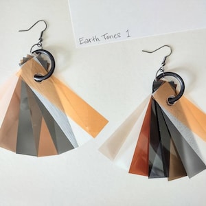 May include: A pair of earrings made from layered translucent paper in shades of brown, orange, and white. The paper is attached to a black metal ring and hook.