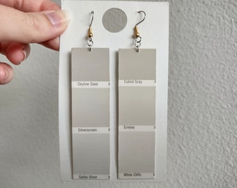 May include: Two pairs of earrings made from paint chip cards. The left earring has the colors Skyline Steel, Silverscreen, and Garbo Silver. The right earring has the colors Cubist Gray, Ermine, and White Cliffs.