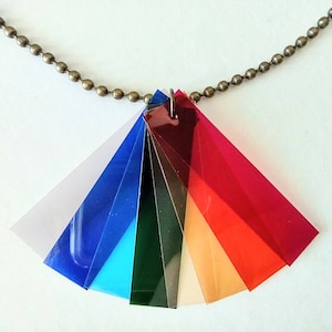 Puede incluir: Un collar con un colgante hecho de múltiples capas de plástico translúcido de colores. El colgante tiene forma de abanico y presenta tonos de rojo, naranja, amarillo, verde, azul y morado.