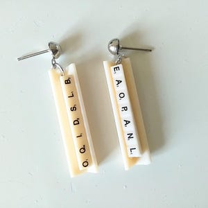 May include: A pair of white dangle earrings with silver hooks. Each earring features a rectangular piece with black letters spelling out "O Q I, D, S, L, B" and "E, A, O, P, A. N. L."