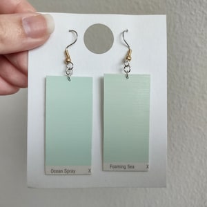 May include: Two pairs of light blue paint chip earrings with silver hooks and gold beads. The paint chips are labeled "Ocean Spray" and "Foaming Sea".