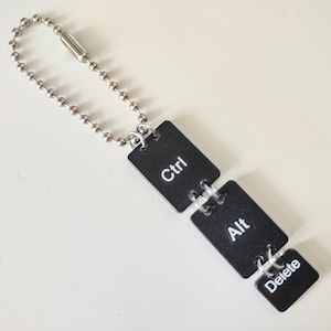 Puede incluir: Un llavero con tres teclas cuadradas negras unidas a una cadena plateada. Las teclas están etiquetadas como "Ctrl", "Alt" y "Suprimir".