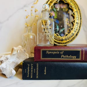 May include: A clear glass bottle labeled "Sulphuric Acid H2 SO4" with a glass stopper, sits atop a stack of books. The books are titled "Synopsis of Pathology" and "The Encyclopedia of Witchcraft and Demonology". A decorative gold framed piece is in the background.