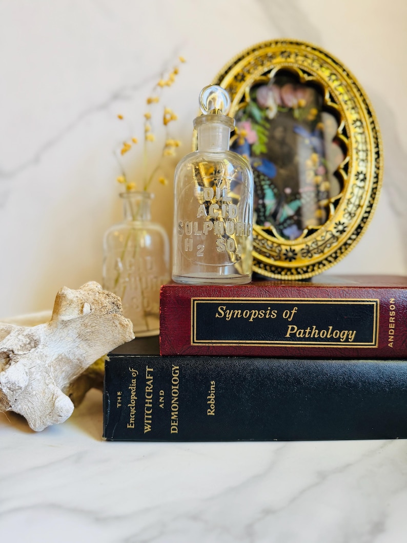 May include: A clear glass bottle labeled "Sulphuric Acid H2 SO4" with a glass stopper, sits atop a stack of books. The books are titled "Synopsis of Pathology" and "The Encyclopedia of Witchcraft and Demonology". A decorative gold framed piece is in the background.