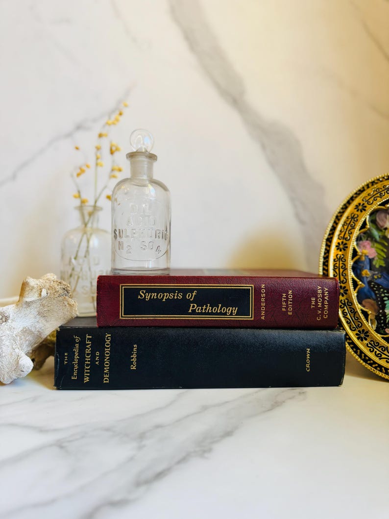 May include: A still life arrangement featuring a clear glass bottle with a stopper, stacked books, and a decorative plate. The books include "Synopsis of Pathology" and "The Encyclopedia of Witchcraft and Demonology."
