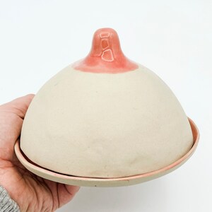 Könnte beinhalten: Eine Keramik-Butterdose in Form einer Brust, mit einer beigefarbenen Kuppel und einer rosa Brustwarze. Die Dose steht auf einem passenden runden Sockel mit rosa Rand. Das Design ist skurril und künstlerisch.