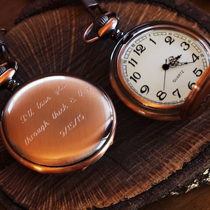 May include: A copper pocket watch with a chain. The watch face has black Roman numeral markings and the words "Quartz" printed on it. The back of the watch is engraved with the message "I'll love you through thick & thin 9/15/19".