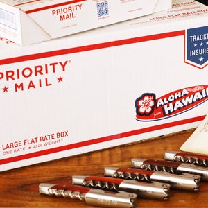 May include: Two white Priority Mail boxes with red and blue accents. The larger box has the text "ALOHA from HAWAII" printed on it.  The smaller box has the text "FLAT RATE ENVELOPE" printed on it.  There are five silver and red corkscrews on a wooden surface.  Two small white fabric bags with the text "The Engraver" printed on them are in the background.