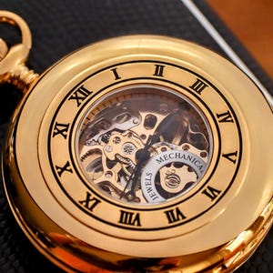 May include: A gold-colored mechanical pocket watch with Roman numeral hour markers. The watch face is visible, showcasing the intricate internal gears and the words "Mechanical" and "Jewels". The watch has a loop at the top for a chain.
