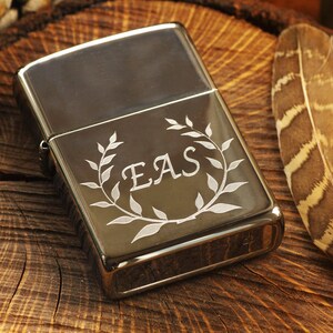 May include: A silver-colored lighter with a decorative leaf wreath and the initials "EAS" engraved on the front. The lighter is set on a rustic wooden surface, with two brown and tan feathers to the right.