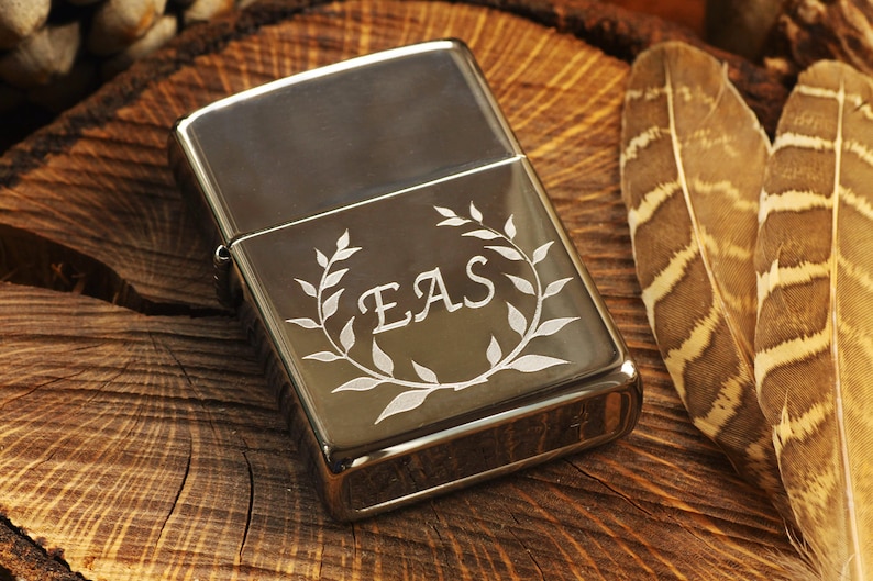May include: A silver-colored lighter with a decorative leaf wreath and the initials "EAS" engraved on the front. The lighter is set on a rustic wooden surface, with two brown and tan feathers to the right.