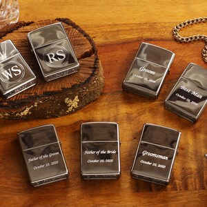 May include: A collection of silver-toned lighters, some engraved with initials and titles like "Groom" and "Best Man," alongside a pocket watch. The lighters are arranged on a wooden surface, suggesting a wedding or celebratory theme.