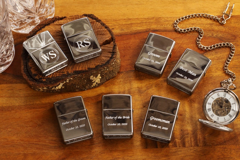 May include: A collection of silver-toned lighters, some engraved with initials and titles like "Groom" and "Best Man," alongside a pocket watch. The lighters are arranged on a wooden surface, suggesting a wedding or celebratory theme.