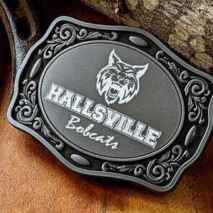 Puede incluir: Una hebilla de cinturón plateada con un diseño ornamentado. La hebilla presenta un óvalo gris con las palabras "Hallsville Bobcats" y una silueta blanca de un lince.