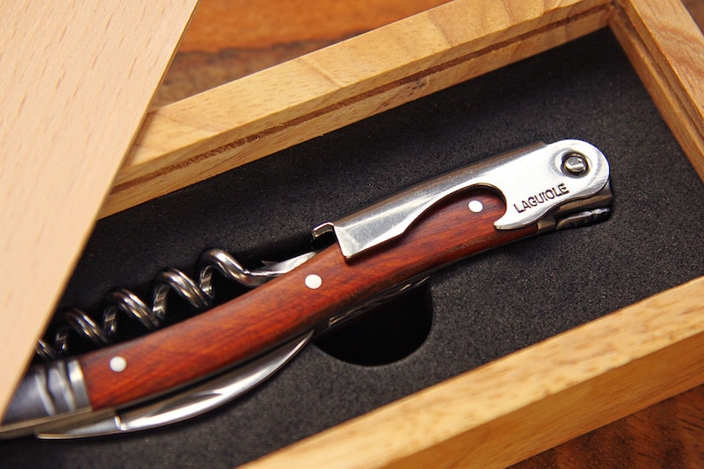 K&ouml;nnte beinhalten: Eine Holzkiste mit einem roten und silbernen Korkenzieher mit einem Holzgriff. Der Korkenzieher hat einen silbernen Flaschen&ouml;ffner und ein kleines Messer. Die Kiste ist ge&ouml;ffnet und der Korkenzieher befindet sich darin. Der Korkenzieher ist mit "LAGUIOLE" beschriftet.