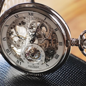 May include: A silver pocket watch with an open face, revealing the intricate inner workings of the mechanism. The watch features Roman numerals and delicate hands, with a chain attached. The watch is resting on a textured black surface.