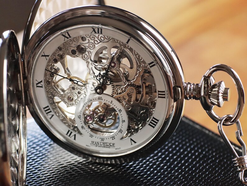 May include: A silver pocket watch with an open face, revealing the intricate inner workings of the mechanism. The watch features Roman numerals and delicate hands, with a chain attached. The watch is resting on a textured black surface.