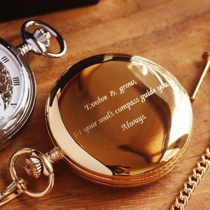 May include: Two pocket watches on a wooden surface. One is silver with a visible mechanism and a white face. The other is gold-toned with the inscription "Evolve & grow, let your soul's compass guide you Always.". A gold chain is attached.