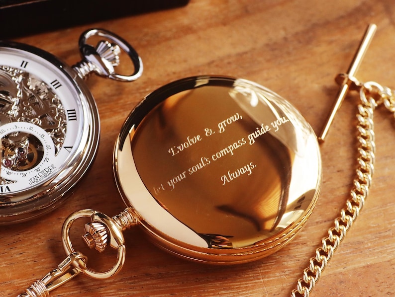 May include: Two pocket watches on a wooden surface. One is silver with a visible mechanism and a white face. The other is gold-toned with the inscription "Evolve & grow, let your soul's compass guide you Always.". A gold chain is attached.