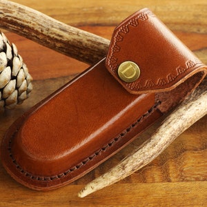 May include: A brown leather knife sheath with a decorative flap and a brass button. The sheath is stitched along the edges and has a curved shape. The background includes pine cones and a deer antler.