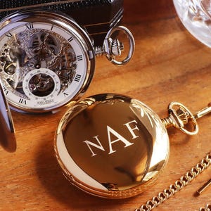 May include: A gold pocket watch with a chain and a second watch with the initials "NAF" engraved on the cover. The open-faced watch reveals the intricate mechanical workings and Roman numeral hour markers. The watches rest on a wooden surface.