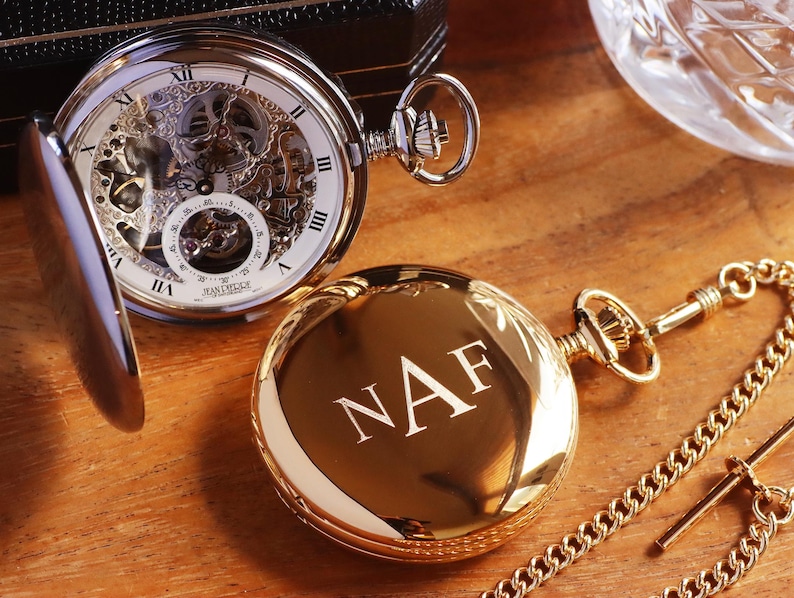 May include: A gold pocket watch with a chain and a second watch with the initials "NAF" engraved on the cover. The open-faced watch reveals the intricate mechanical workings and Roman numeral hour markers. The watches rest on a wooden surface.