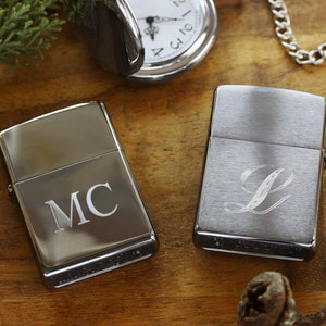 May include: Two silver lighters with personalized engravings. One lighter features the initials "MC", while the other displays the letter "L". A pocket watch and chain are visible in the background, along with greenery.