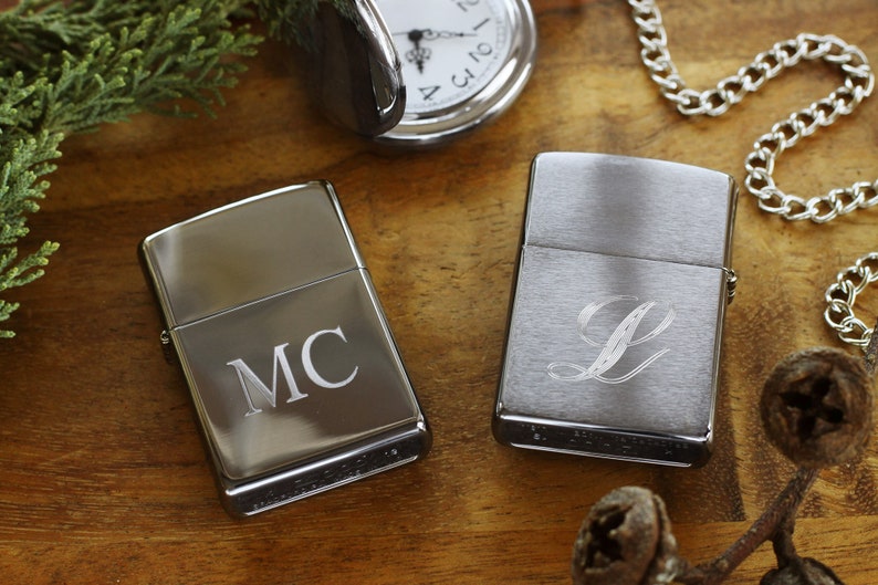 May include: Two silver lighters with personalized engravings. One lighter features the initials "MC", while the other displays the letter "L". A pocket watch and chain are visible in the background, along with greenery.