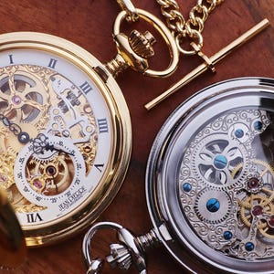 May include: Two antique pocket watches with intricate mechanical movements. One watch is gold-toned with a white face and Roman numerals. The other is silver-toned, showcasing the inner workings. Both have chains and ornate details.