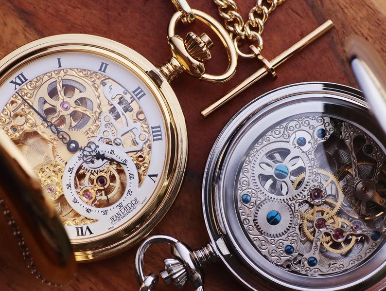 May include: Two antique pocket watches with intricate mechanical movements. One watch is gold-toned with a white face and Roman numerals. The other is silver-toned, showcasing the inner workings. Both have chains and ornate details.