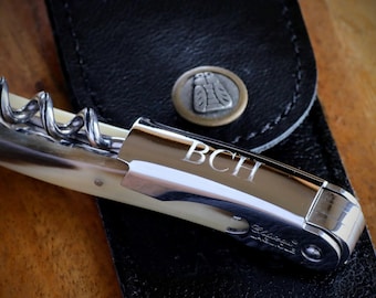 May include: A close-up of a silver and ivory corkscrew with the initials "BCH" engraved on the handle. The corkscrew is partially open, revealing the spiral. It rests on a black leather case with a decorative metal button.
