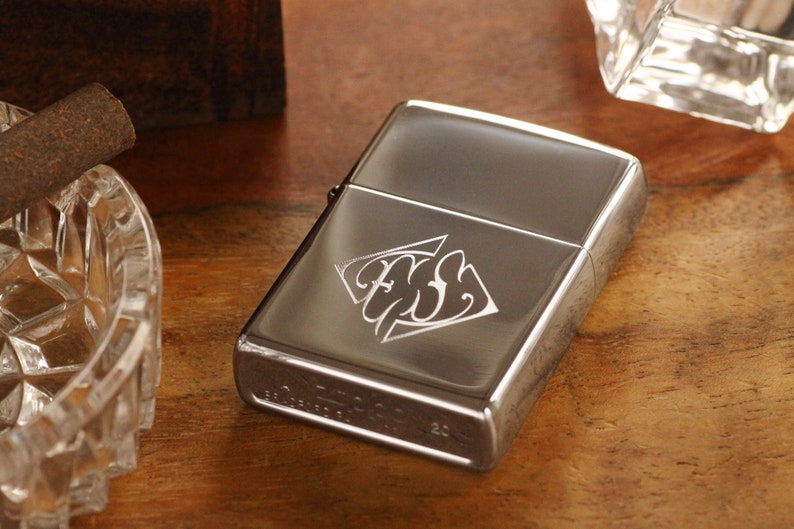 May include: A silver-colored lighter with an engraved design on the front. The lighter is rectangular and has a shiny finish. A cigar and a crystal ashtray are also visible on a wooden surface.
