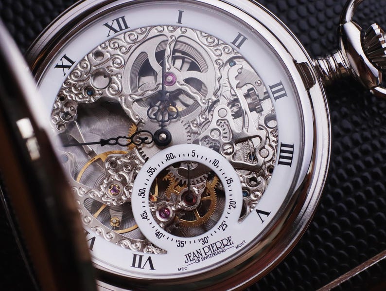 May include: Close-up of a silver Jean Pierre of Switzerland pocket watch with a white face and Roman numerals. The watch features an intricate open-face design, showcasing the internal gears and mechanisms. The watch has a small seconds dial and is set against a dark background.