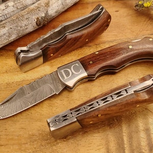 May include: Three folding knives with wooden handles and patterned steel blades are displayed on a wooden surface. The central knife is open, revealing the blade's intricate design and the initials "DC" engraved on the bolster. The handles are a rich brown, accented with brass rivets.