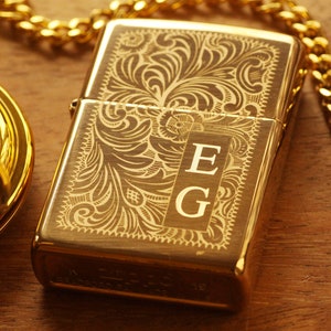 May include: A gold-colored Zippo lighter with an engraved floral design and the initials "EG". A gold pocket watch and chain are visible in the background, suggesting a vintage or luxury theme.