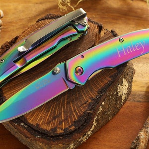 May include: Two rainbow-colored pocket knives with a silver clip. The knife on the right has the name "Haley" engraved on the blade.