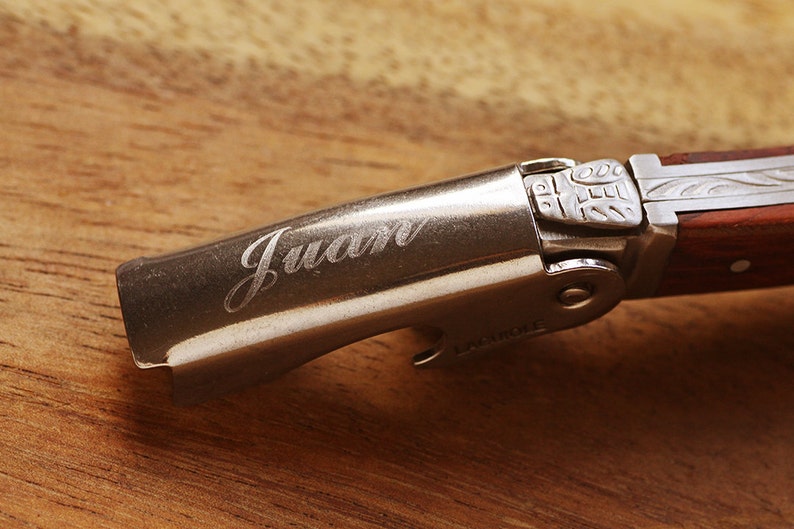 K&ouml;nnte beinhalten: Ein silberner Flaschen&ouml;ffner mit einem Holzgriff. Der &Ouml;ffner hat ein dekoratives Design auf dem Griff und das Wort "Juan" ist in den Metallteil graviert.