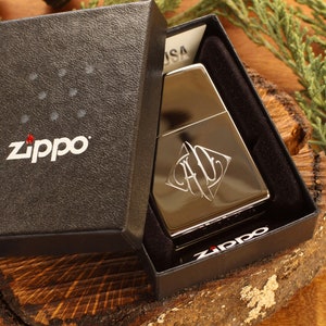 May include: A silver Zippo lighter with an engraved design, presented in a black gift box. The box features the Zippo logo in white. The lighter is displayed on a wooden surface with green foliage in the background.