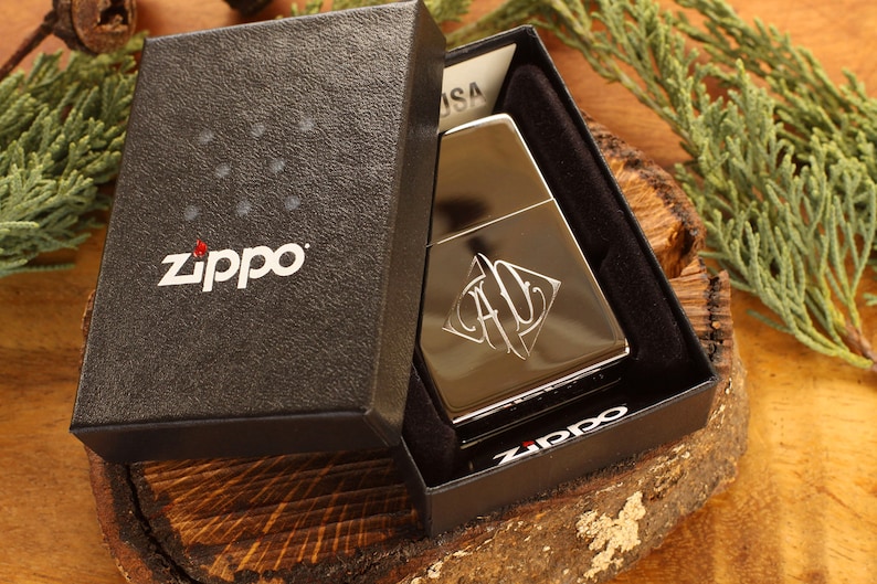 May include: A silver Zippo lighter with an engraved design, presented in a black gift box. The box features the Zippo logo in white. The lighter is displayed on a wooden surface with green foliage in the background.