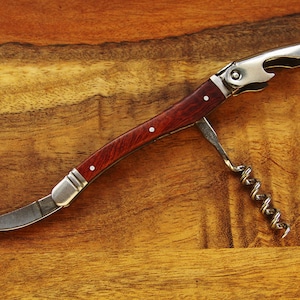 K&ouml;nnte beinhalten: Ein silberner und h&ouml;lzerner Flaschen&ouml;ffner mit einem Korkenzieher, einem Flaschen&ouml;ffner und einem kleinen Messer. Der &Ouml;ffner liegt auf einer Holzoberfl&auml;che.