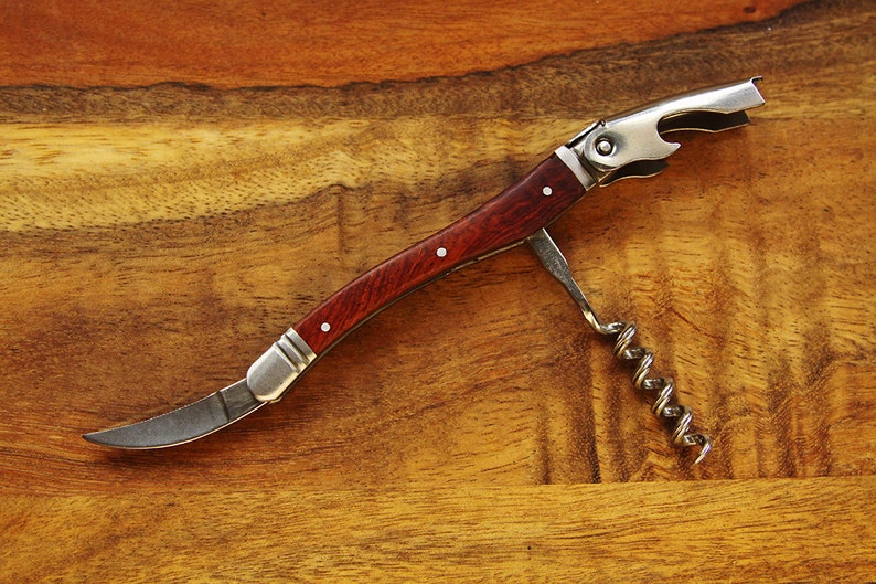 K&ouml;nnte beinhalten: Ein silberner und h&ouml;lzerner Flaschen&ouml;ffner mit einem Korkenzieher, einem Flaschen&ouml;ffner und einem kleinen Messer. Der &Ouml;ffner liegt auf einer Holzoberfl&auml;che.