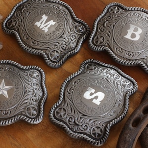 May include: Four silver-toned belt buckles with ornate designs. Each buckle features a star and a different letter or number in the center. The buckles are arranged on a wooden surface.