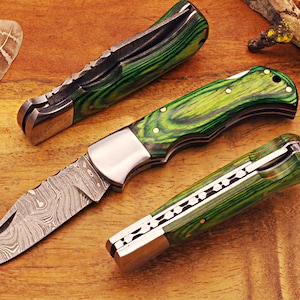 May include: Three folding knives with green and black patterned handles and silver blades are displayed on a wooden surface. The blades have a unique pattern. The handles have a wood grain appearance. The knives are open and closed.