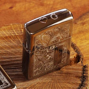 May include: A silver lighter with an engraved floral design and the letter "G" on the top. The lighter has a heart symbol next to the letter.