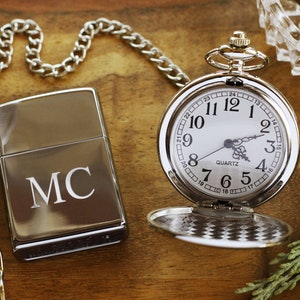 May include: A silver pocket watch with a white face and a matching silver lighter engraved with the initials "MC" sit on a wooden surface. A silver chain and a glass ashtray are also visible.