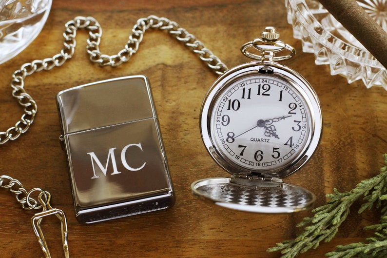 May include: A silver pocket watch with a white face and a matching silver lighter engraved with the initials "MC" sit on a wooden surface. A silver chain and a glass ashtray are also visible.