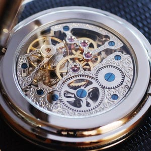 May include: A silver pocket watch with a transparent face, showcasing the intricate gold and silver gears and jewels of the watch mechanism. The watch has a silver chain and a hinged cover. The watch is a classic timepiece.